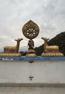 Golden Buddhist statues with deer and wheel of Dharma on a rooftop, mountains in background, cloudy sky.